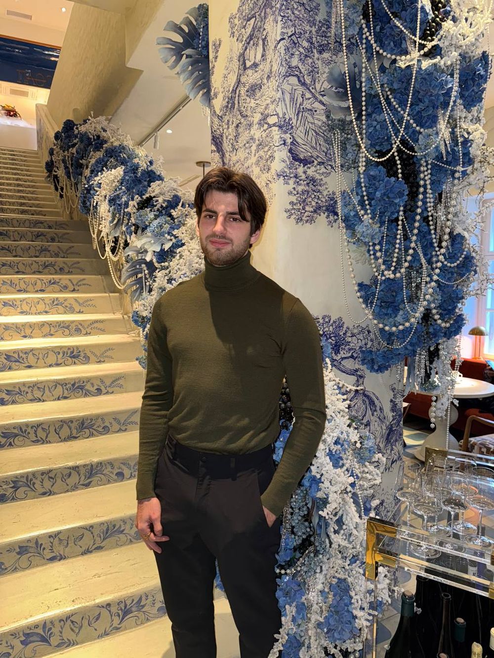 Alexis Dubain, pull vert olive, dans un escalier décoré de fleurs bleues
